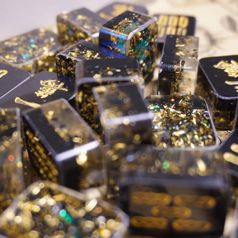 Op de afbeelding: Een verzameling zwarte en heldere harsblokken met gouden vlokken en gouden letters. De blokken zijn rechthoekig en hebben een glanzende afwerking. Sommige blokken hebben gouden Chinese karakters.