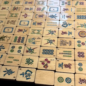 Puede incluir: Primer plano de un juego de fichas de Mahjong de madera, que muestra una variedad de s&iacute;mbolos y caracteres en diferentes colores. Las fichas est&aacute;n dispuestas en una cuadr&iacute;cula, destacando los dise&ntilde;os detallados y los elementos del juego. Algunas fichas tienen el texto "Tiley By Tash".