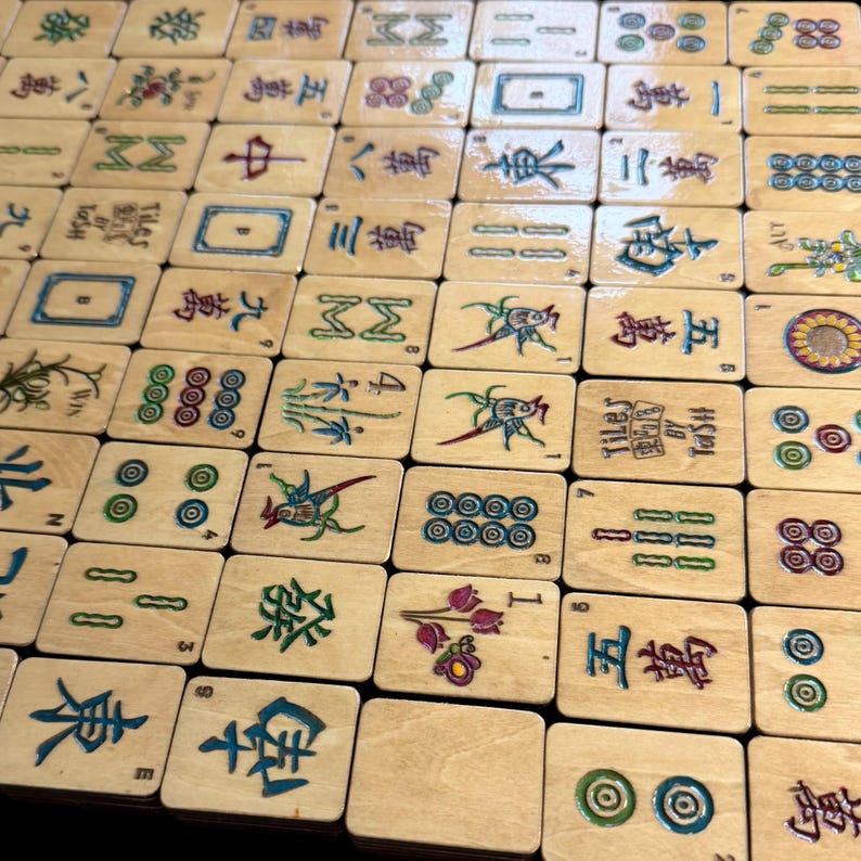 Puede incluir: Primer plano de un juego de fichas de Mahjong de madera, que muestra una variedad de s&iacute;mbolos y caracteres en diferentes colores. Las fichas est&aacute;n dispuestas en una cuadr&iacute;cula, destacando los dise&ntilde;os detallados y los elementos del juego. Algunas fichas tienen el texto "Tiley By Tash".