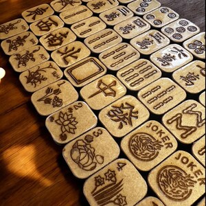 May include: A collection of wooden Mahjong tiles with engraved symbols and characters. The tiles are beige with dark brown engravings, including numbers, suits, and special tiles like "Joker". The tiles are arranged on a dark wooden surface.