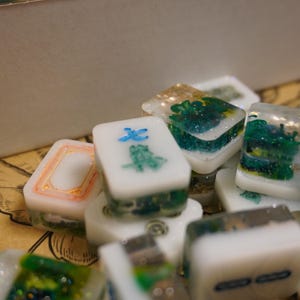 May include: Close-up of several white and green Mahjong tiles. Some tiles feature blue and green symbols, while others have decorative green and gold designs. The tiles are stacked, showcasing their intricate details.