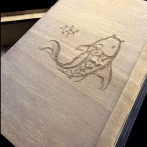 May include: Wooden box with a detailed etched design of a koi fish and a Japanese character. The fish is centered on the lid, with the character above it. The wood has a natural, light brown color.