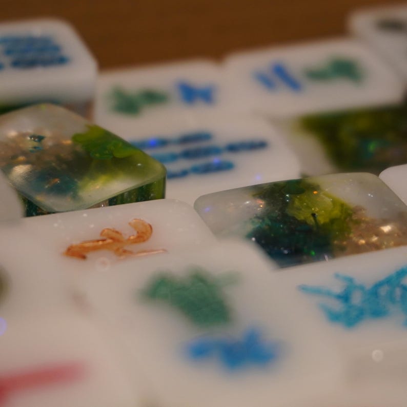 May include: Close-up of several white resin tiles with embedded designs. Some tiles feature green and blue glitter, while others have orange or blue shapes. The tiles are slightly translucent, with a blurred background.