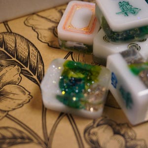 May include: Close-up of several white and clear resin Mahjong tiles with green, gold, and glitter accents. Some tiles have Chinese characters. The tiles are on a brown floral patterned surface.