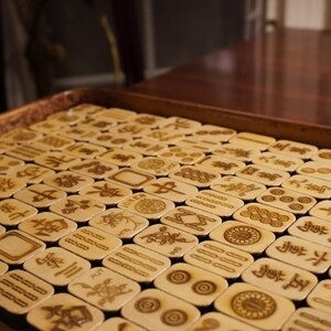 May include: A full tray of ivory-colored Mahjong tiles, each engraved with symbols and characters in brown. The tiles are arranged neatly, filling the frame. Some tiles feature the word "JOKER" and other symbols.
