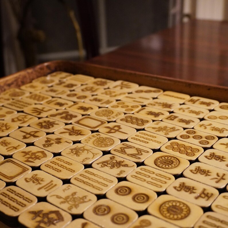 May include: A full tray of ivory-colored Mahjong tiles, each engraved with symbols and characters in brown. The tiles are arranged neatly, filling the frame. Some tiles feature the word "JOKER" and other symbols.