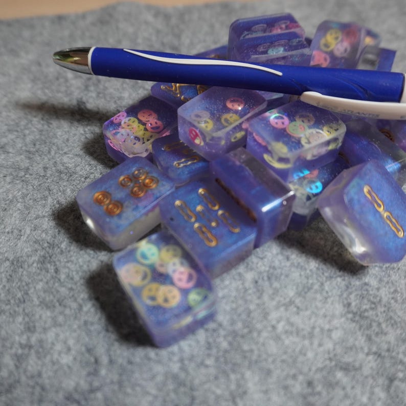 May include: A collection of small, translucent blue resin blocks with embedded decorative elements, including gold-colored shapes and colorful smiley faces. A blue pen rests on top of the blocks. The blocks are scattered on a gray surface.