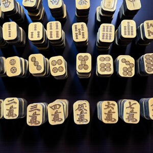 May include: A collection of wooden Mahjong tiles, each with unique symbols and characters. The tiles are stacked in rows, showcasing the variety of designs and the game's components. The wood has a light color with dark edges, creating a contrast.