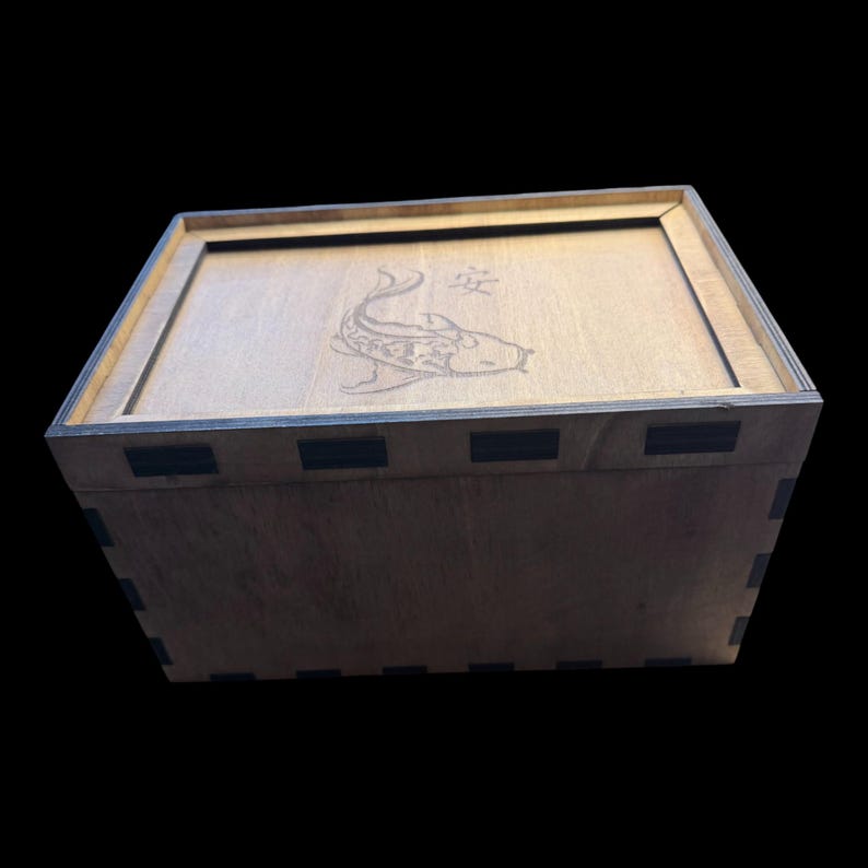 May include: A wooden box with a lid featuring a laser-etched koi fish design and Japanese characters. The box is constructed from light-colored wood with dark accents at the corners and edges. The box is rectangular and has a simple, clean design.