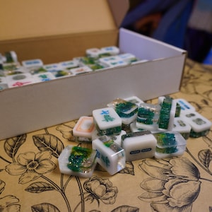 May include: A collection of white and clear resin Mahjong tiles with green and blue accents, some with Chinese characters. The tiles are scattered on a floral patterned surface, with a cardboard box in the background.