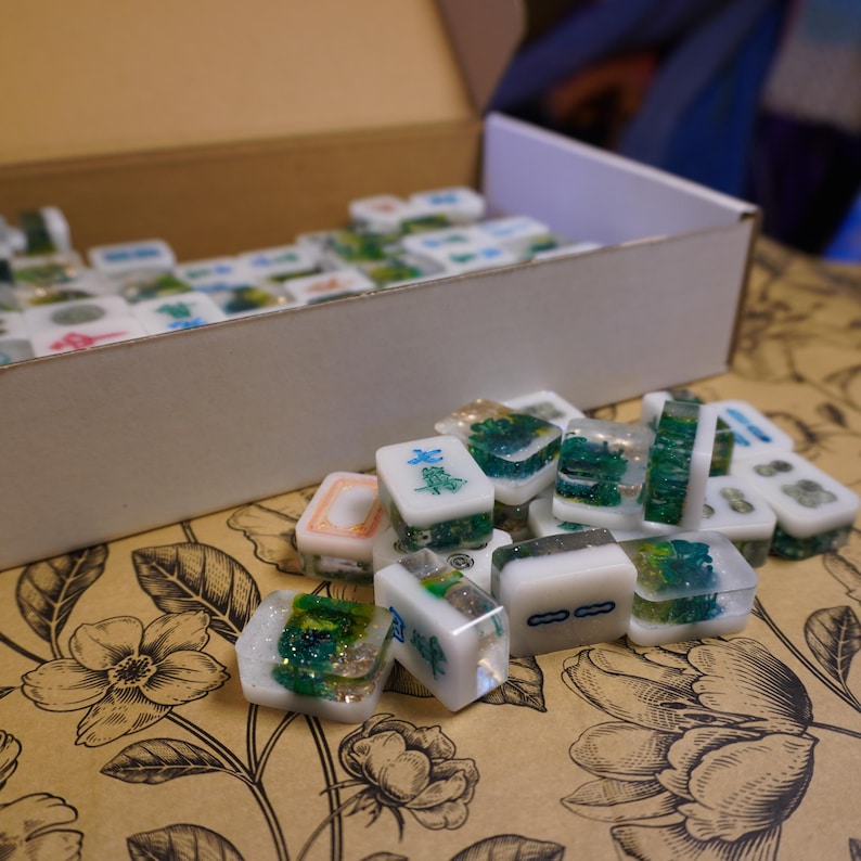 May include: A collection of white and clear resin Mahjong tiles with green and blue accents, some with Chinese characters. The tiles are scattered on a floral patterned surface, with a cardboard box in the background.