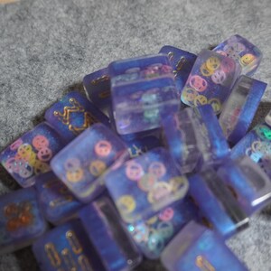 May include: A collection of small, rectangular resin pieces in shades of blue and purple. Some pieces feature gold designs, while others have colorful smiley face patterns. The pieces are scattered on a gray surface.