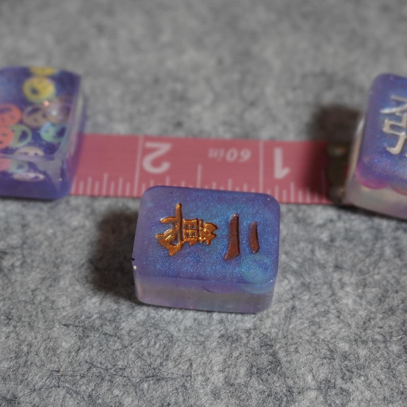 May include: Close-up of three small, square, purple resin objects. The central object has gold Chinese characters. A pink measuring tape with inch markings is in the background. The objects are on a gray surface.
