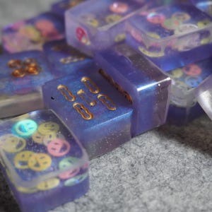 May include: Close-up of several rectangular resin pieces in shades of purple and blue. Some pieces have gold-colored embellishments, while others contain colorful smiley face shapes. The pieces are scattered on a gray surface.