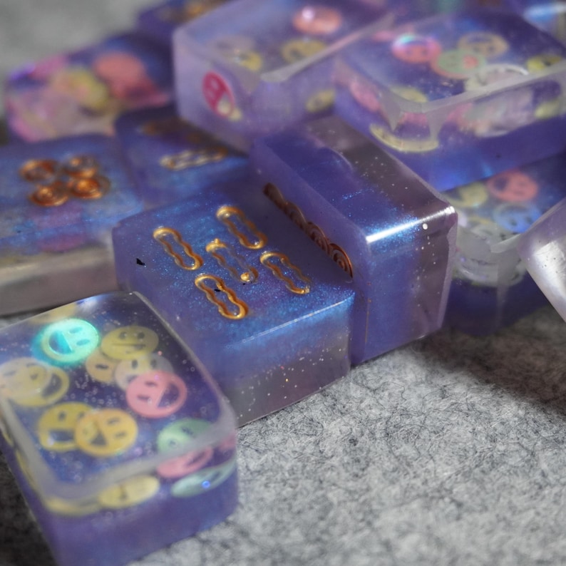May include: Close-up of several rectangular resin pieces in shades of purple and blue. Some pieces have gold-colored embellishments, while others contain colorful smiley face shapes. The pieces are scattered on a gray surface.