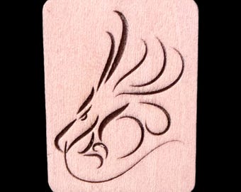 May include: A rectangular, pale pink wooden plaque with rounded corners. A dark brown dragon head design is etched into the wood. The dragon's head is in profile, with curved lines forming the mane and body.
