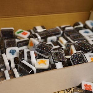 May include: A collection of colorful Mahjong tiles in a cardboard box. The tiles feature black glitter, white, and black stripes, and various symbols in orange, green, and blue. The game pieces are rectangular and have a glossy finish.