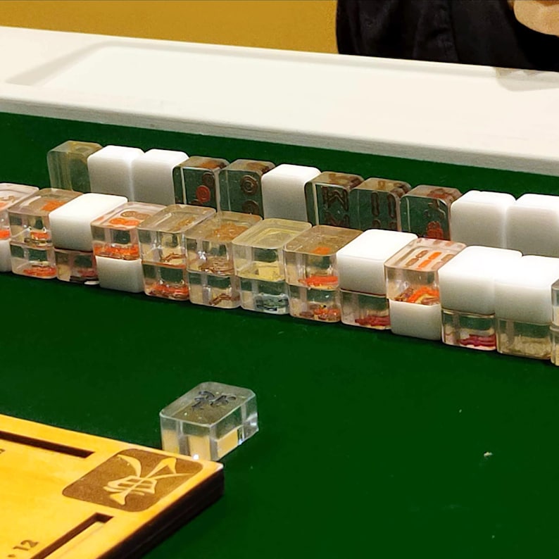 May include: A collection of Mahjong tiles, some clear with orange and red details, others white or green. The tiles are arranged on a green surface, with a wooden game board visible in the foreground. The game board has a gold emblem.