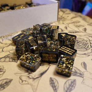 Op de afbeelding: Een verzameling zwarte harsdobbelstenen met gouden en groene glitterinsluitsels. De dobbelstenen hebben verschillende gouden markeringen, waaronder stippen en lijnen. Ze liggen op een oppervlak met bloemenpatroon, sommige vallen uit een kartonnen doos.