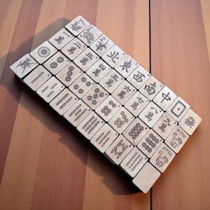 May include: A collection of wooden Mahjong tiles, each displaying engraved Chinese characters and symbols, including dots, bamboo, and dragons. The tiles are arranged diagonally on a brown surface. Some tiles are marked with the word "Joker".