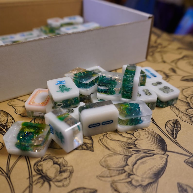 May include: A collection of handmade Mahjong tiles. The tiles are white with green and gold accents, and some feature Chinese characters. They are arranged on a floral patterned surface, with a box in the background.