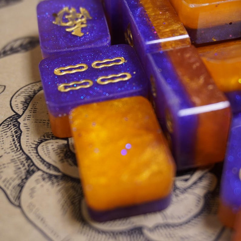Pu&ograve; includere: Primo piano di diverse tessere di gioco in resina viola e arancione. Le tessere hanno dettagli dorati e glitter. Alcune tessere hanno simboli e segni dorati. Le tessere sono disposte su una superficie a motivi.