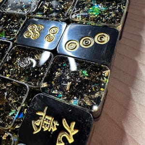 Op de afbeelding: Close-up van een verzameling rechthoekige hars tegels. Sommige tegels zijn zwart met gouden accenten en Chinese karakters. Andere zijn zilver met gouden cirkelvormige ontwerpen. De tegels zijn gevuld met gouden en iriserende vlokken.