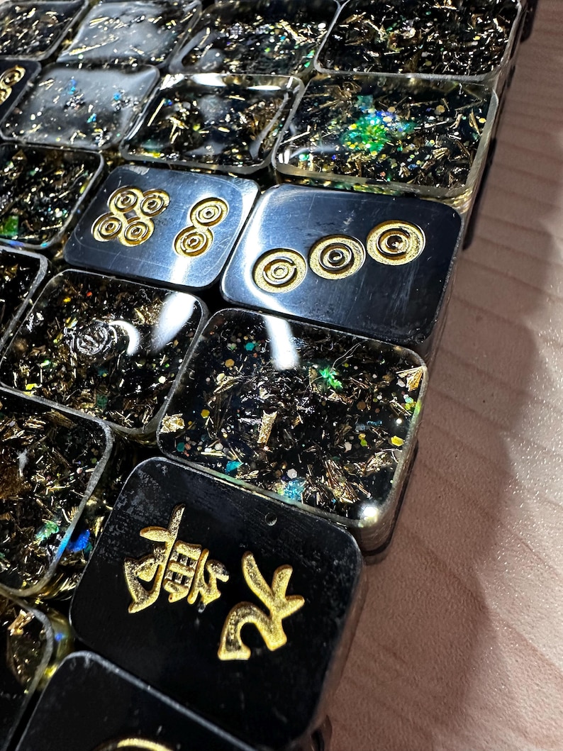 Op de afbeelding: Close-up van een verzameling rechthoekige hars tegels. Sommige tegels zijn zwart met gouden accenten en Chinese karakters. Andere zijn zilver met gouden cirkelvormige ontwerpen. De tegels zijn gevuld met gouden en iriserende vlokken.