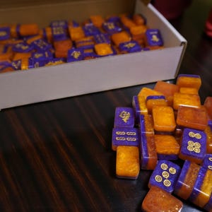 May include: A collection of orange and purple Mahjong tiles with gold detailing. Some tiles are stacked, while others are in a white box. The tiles are on a dark wooden surface.