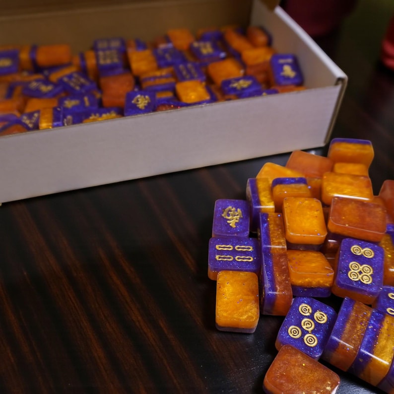 May include: A collection of orange and purple Mahjong tiles with gold detailing. Some tiles are stacked, while others are in a white box. The tiles are on a dark wooden surface.