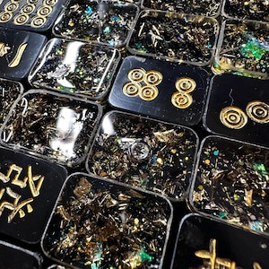 Pu&ograve; includere: Primo piano di pi&ugrave; piastrelle rettangolari nere in resina con accenti dorati e foglia d'oro e glitter incorporati. Alcune piastrelle presentano simboli o motivi dorati. Le piastrelle sono disposte vicine, creando un effetto a mosaico.