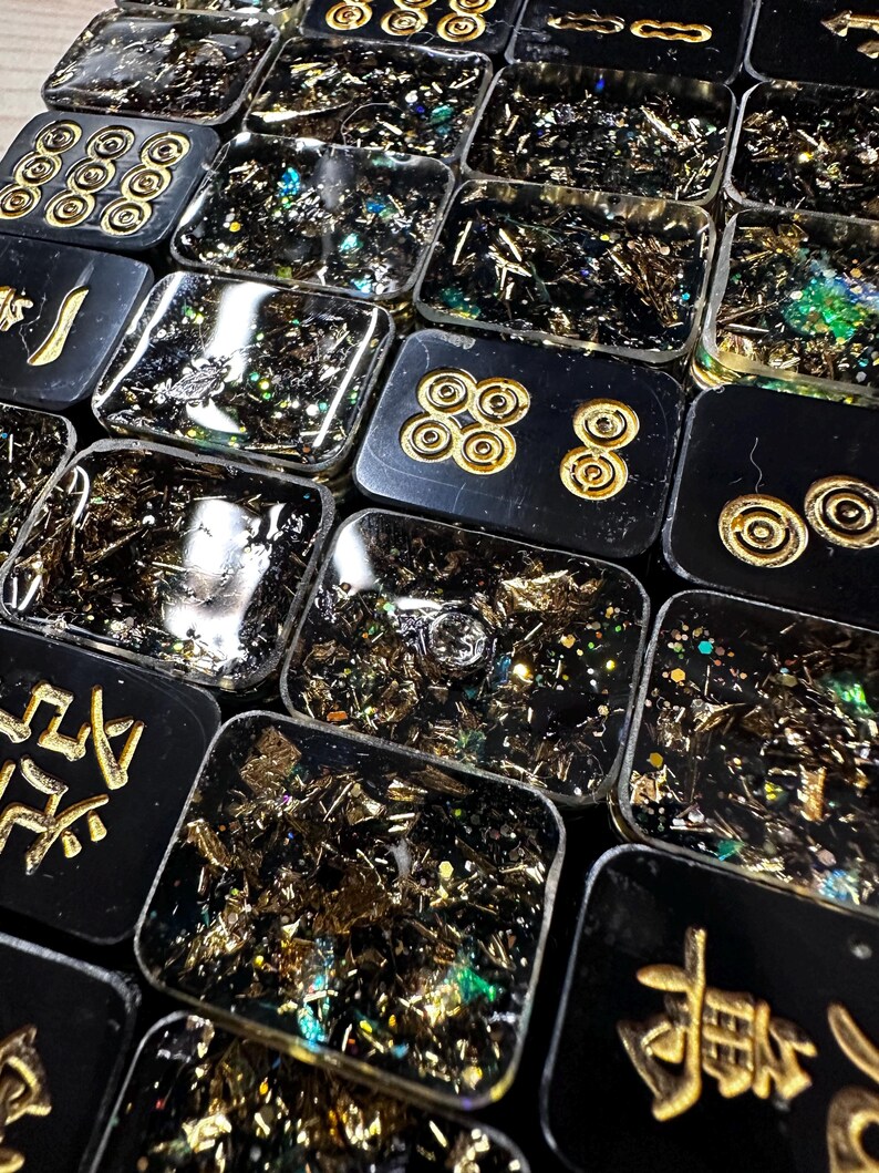 Pu&ograve; includere: Primo piano di pi&ugrave; piastrelle rettangolari nere in resina con accenti dorati e foglia d'oro e glitter incorporati. Alcune piastrelle presentano simboli o motivi dorati. Le piastrelle sono disposte vicine, creando un effetto a mosaico.