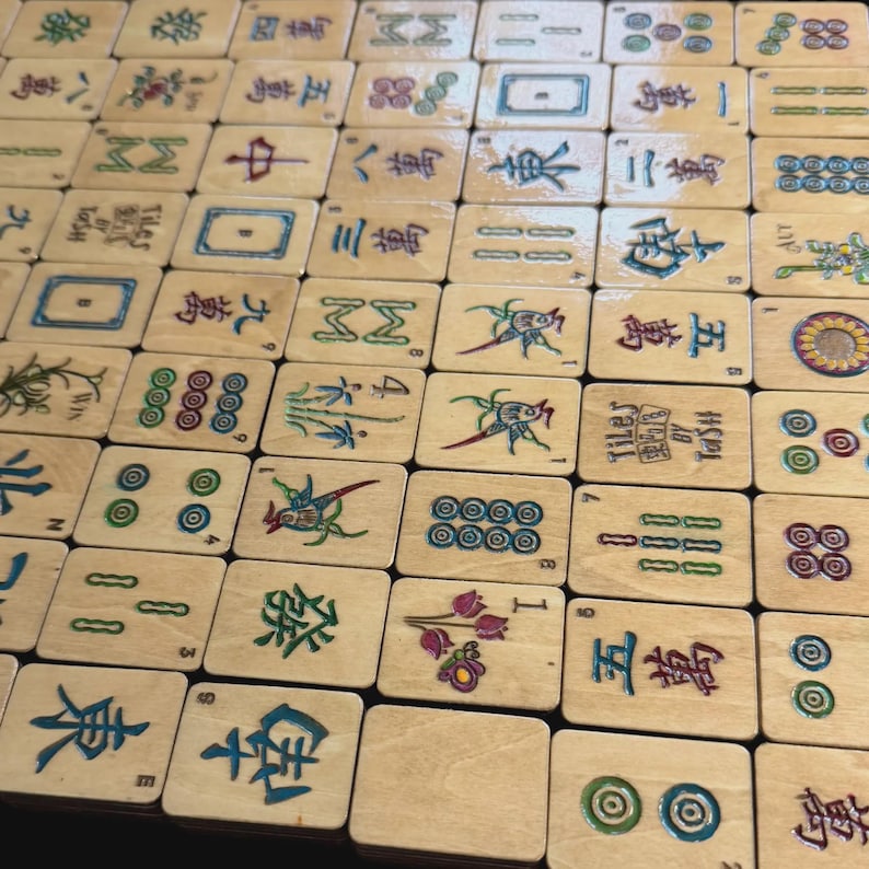 May include: A set of wooden Mahjong tiles, each displaying distinct symbols and characters in various colours. The tiles are arranged in a grid, showcasing the diverse designs and numbers. Some tiles feature text, including "Tiles by TaSH".