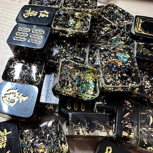 Op de afbeelding: Een verzameling zwarte harsblokken met goudkleurige Chinese karakters en symbolen. De blokken zijn gevuld met goud en iriserende glitter, wat een sprankelend effect cre&euml;ert. De blokken zijn rechthoekig en vierkant.
