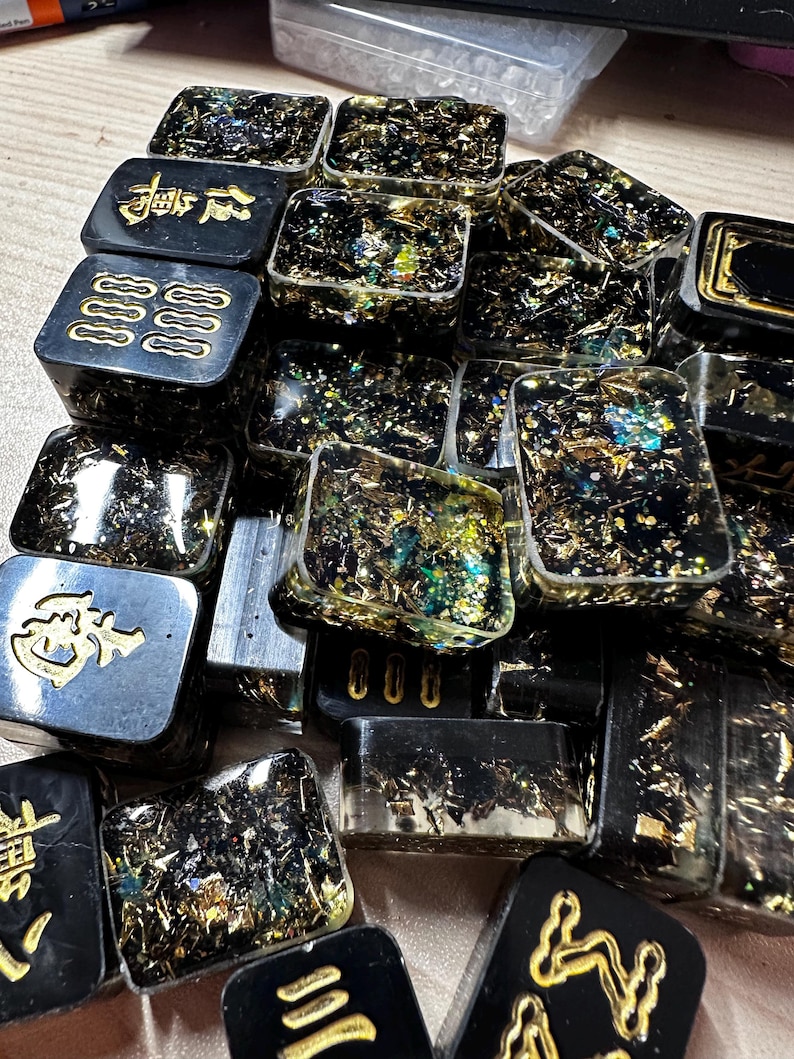 Op de afbeelding: Een verzameling zwarte harsblokken met goudkleurige Chinese karakters en symbolen. De blokken zijn gevuld met goud en iriserende glitter, wat een sprankelend effect cre&euml;ert. De blokken zijn rechthoekig en vierkant.
