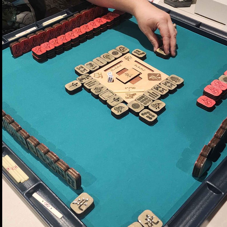May include: A Mahjong game in progress, featuring a teal playing surface. The game includes wooden tiles with symbols, red tiles, and a wooden board. A hand is placing a tile on the board. The game is set up for play.