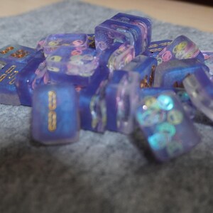 May include: A collection of translucent, iridescent purple resin dice with gold accents and colorful embedded shapes. The dice are scattered on a gray surface, showcasing their unique, decorative design.