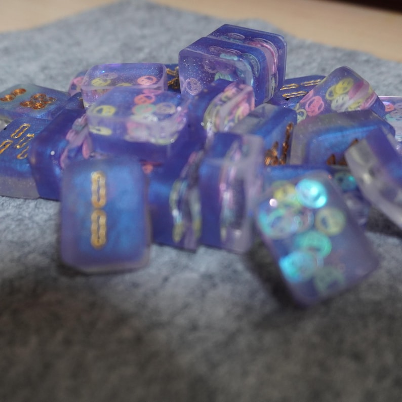May include: A collection of translucent, iridescent purple resin dice with gold accents and colorful embedded shapes. The dice are scattered on a gray surface, showcasing their unique, decorative design.