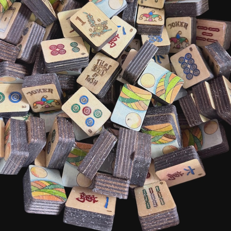 May include: A pile of vintage mahjong tiles, displaying various symbols, numbers, and colourful illustrations. The tiles are made of a light-coloured material with dark brown edges. Some tiles display the word "Joker" and the text "Tiles by Tash".
