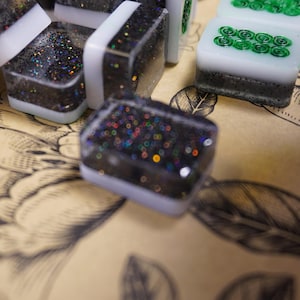 May include: Close-up of several square game tiles. The tiles have a black base with iridescent glitter, white borders, and some have green swirl designs. The tiles are on a tan surface with a floral pattern.