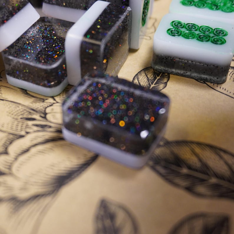 May include: Close-up of several square game tiles. The tiles have a black base with iridescent glitter, white borders, and some have green swirl designs. The tiles are on a tan surface with a floral pattern.