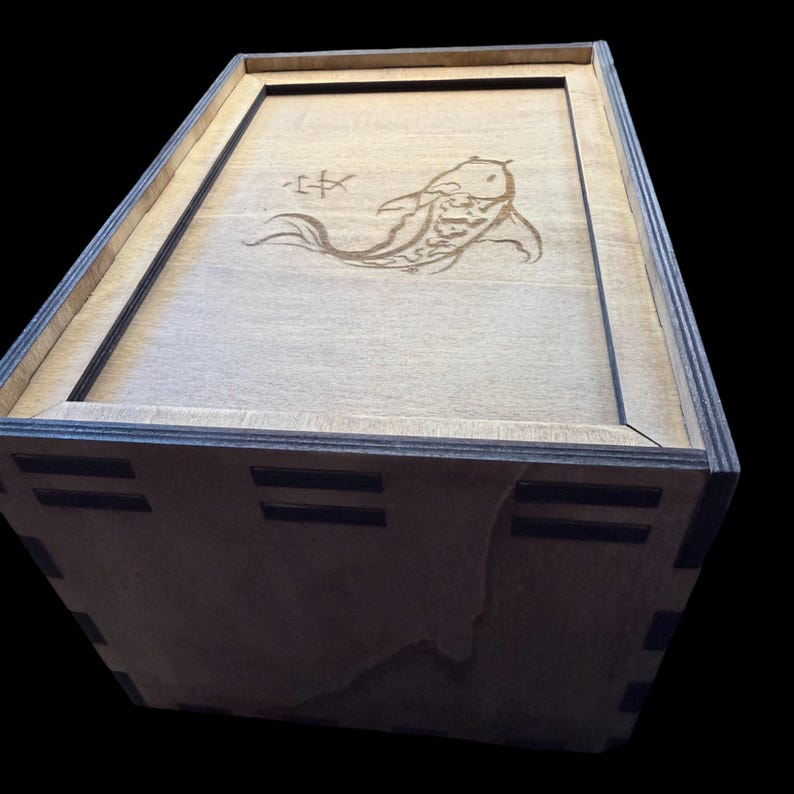 May include: Wooden box with a lid featuring a laser-etched koi fish design. The box is rectangular with a dark border and a light wood finish. The koi fish is depicted in a flowing, stylized manner, with Japanese characters above it.