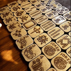 Op de afbeelding: Een verzameling houten Mahjong-tegels met gegraveerde symbolen en karakters. De beige tegels hebben donkerbruine ge&euml;tste ontwerpen, waaronder cijfers, pakken en speciale tegels zoals "Joker". De tegels liggen op een donker houten oppervlak.