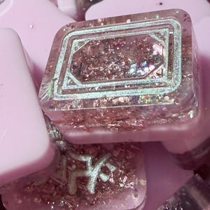 Pu&ograve; includere: Primo piano di un tasto quadrato in resina trasparente con un design geometrico argentato. Il tasto &egrave; riempito di glitter rosa e piccoli fiocchi iridescenti. Altri tasti rosa sono visibili sullo sfondo.
