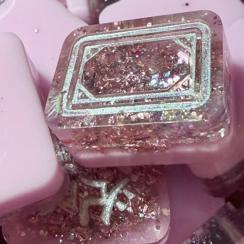 Pu&ograve; includere: Primo piano di un tasto quadrato in resina trasparente con un design geometrico argentato. Il tasto &egrave; riempito di glitter rosa e piccoli fiocchi iridescenti. Altri tasti rosa sono visibili sullo sfondo.