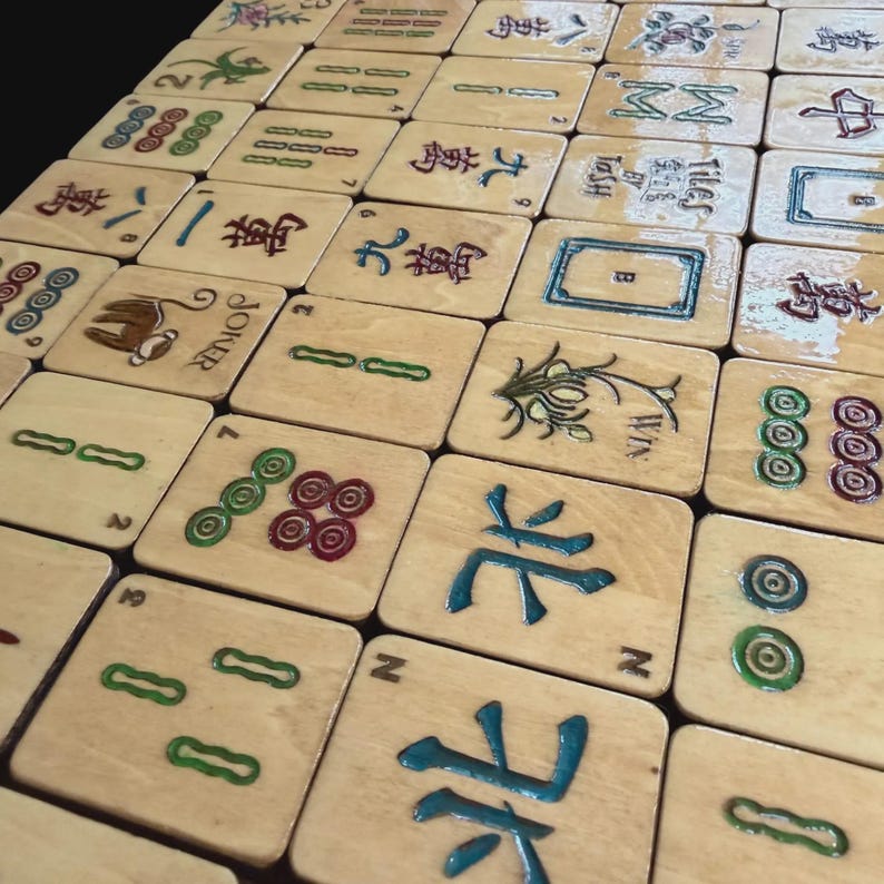 May include: A selection of wooden Mahjong tiles, each displaying unique symbols and characters in shades of green, red, and blue. Some tiles feature numbers, while others showcase detailed designs and the word "JOKER". The tiles are arranged on a light-coloured surface.