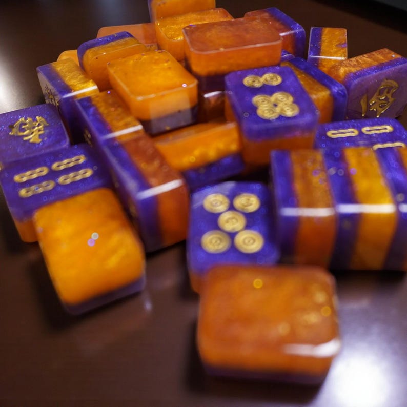 May include: A collection of colorful mahjong tiles in shades of orange and purple. The tiles feature gold symbols and designs, creating a visually appealing and intricate pattern. The tiles are arranged in a pile, showcasing their unique colors and details.