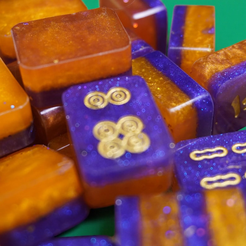 May include: Close-up of a collection of handmade resin game pieces. The pieces are a mix of rectangular and square shapes, with a two-tone color scheme of orange and purple. Some pieces feature gold-colored symbols, and glitter is embedded throughout.