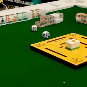 May include: A close-up of a Mahjong set on a green surface. The set includes white tiles with various symbols, clear dice, and a yellow wooden board. The tiles are arranged in rows, with some in clear plastic containers.