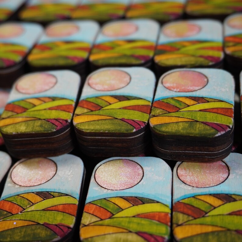 Handmade wooden Mahjong tiles featuring a Sunrise back design with rolling hills and a rising sun. Crafted from layered basswood, precision laser-engraved, and sealed with protective gloss. Available in Riichi or American Mahjong sets, with three finish options: engraved-only, spot color accent on the sun, or fully hand-painted vibrant design. A radiant, artisan Mahjong set inspired by new beginnings.
